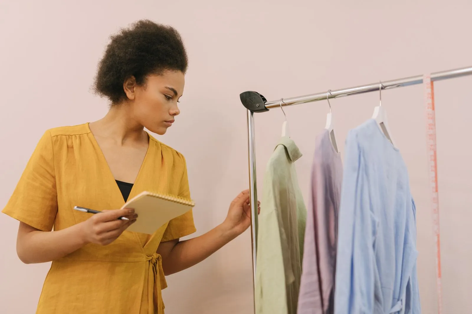 A woman in a mustard linen dress holds an open notepad and pen while working through three garments hanging on a chrome clothing rail against a pale wall.