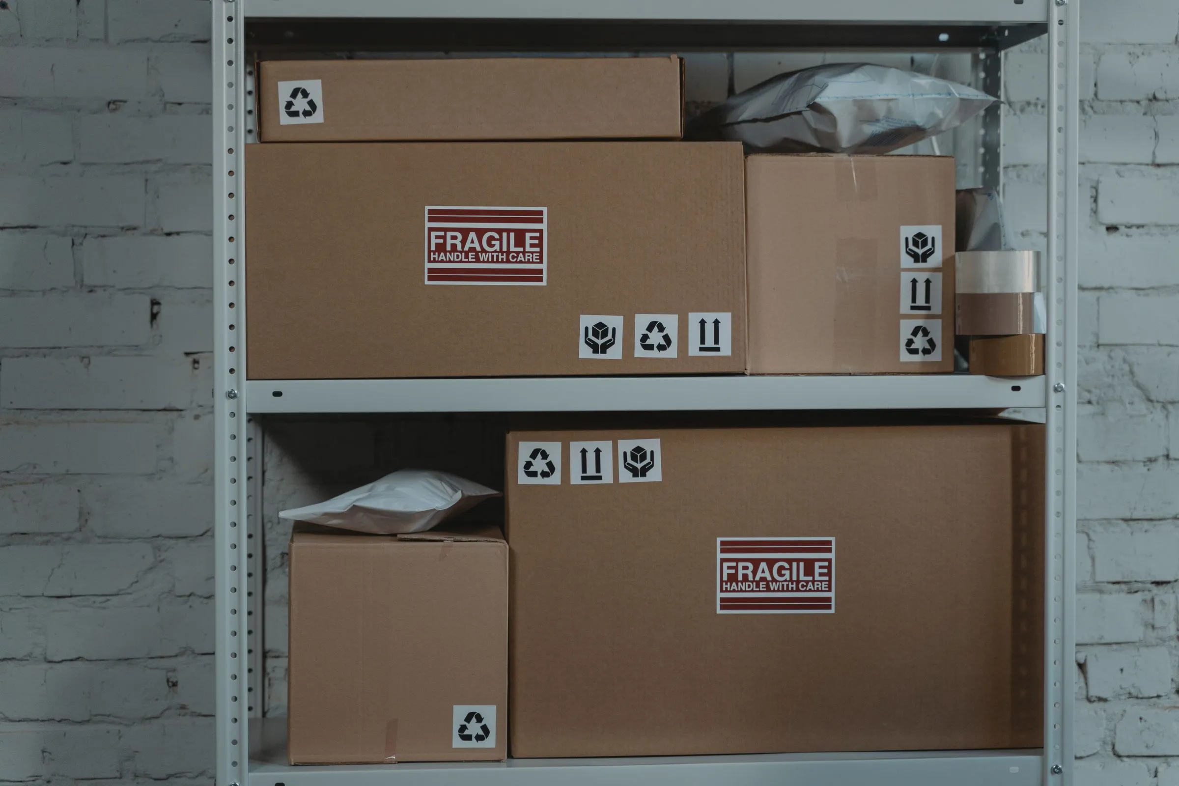 Two shelves of a white steel storage rack holding flat and square kraft cardboard cartons marked &ldquo;Fragile: handle with care&rdquo;, alongside white poly mailer bags and a roll of packing tape, against a whitewashed brick wall.